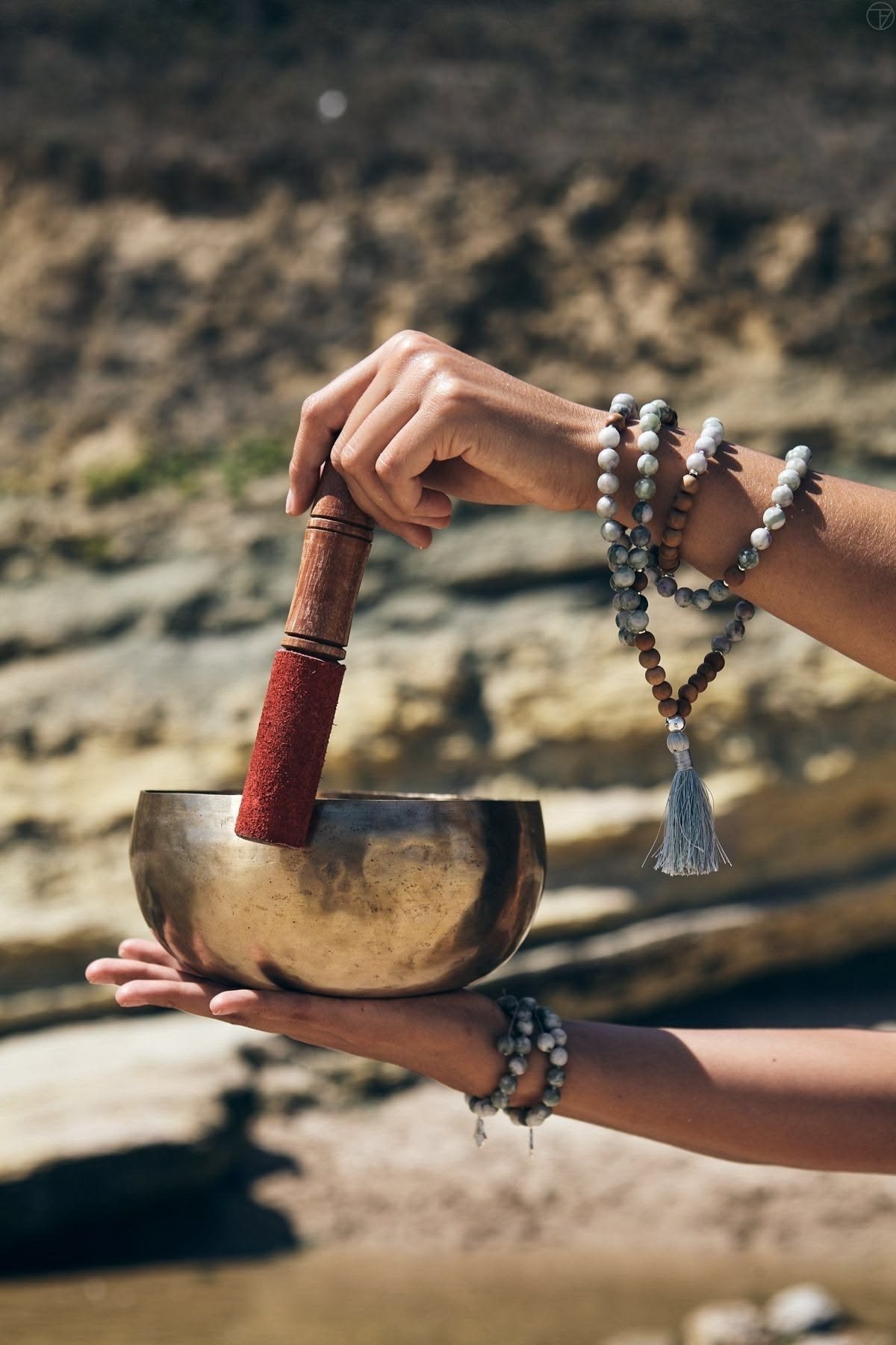 Hands holding a brass singing bowl and mallet, a mala bracelet at the wrist
