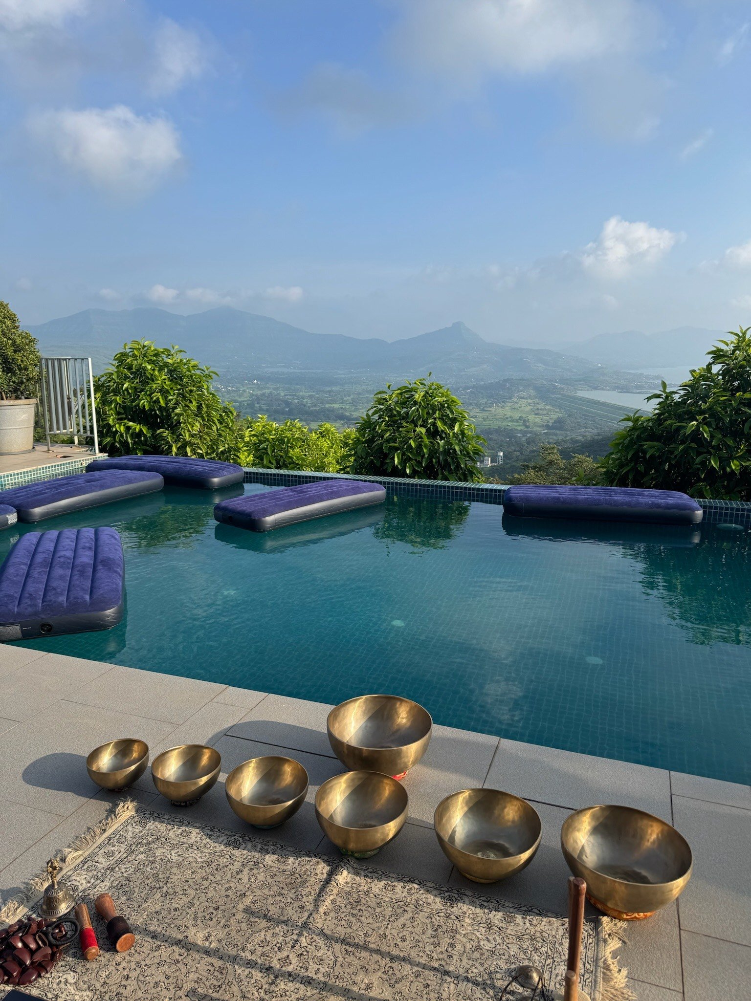 Brass singing bowls laid out on the deck of a hilltop infinity pool, floating beds on the water and green hills beyond