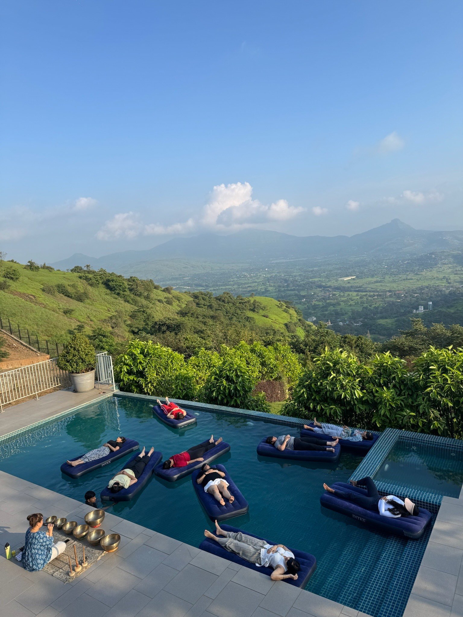 Guests resting on floating beds in a hilltop infinity pool while a practitioner plays singing bowls at the water's edge