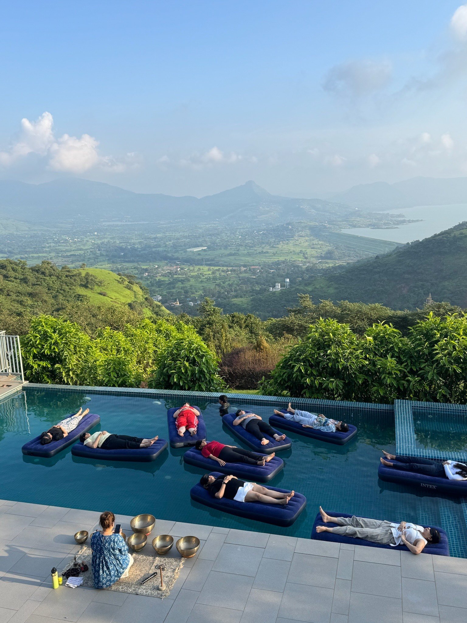 A floating sound bath on an infinity pool high above green hills and a lake, guests resting on floating beds