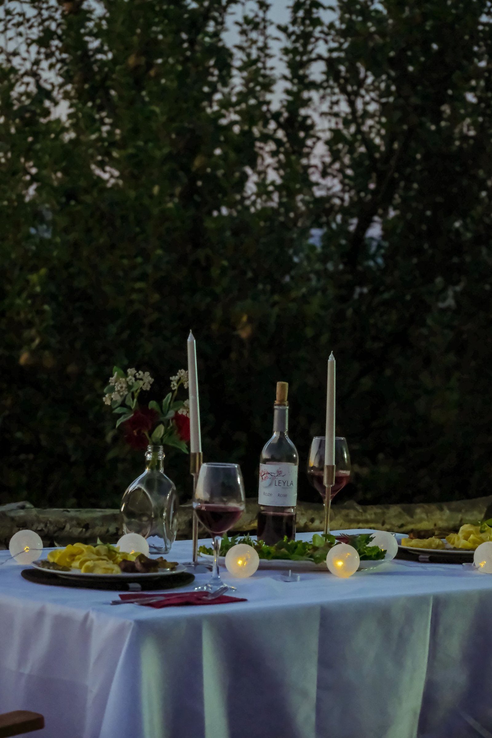 A garden dinner table set with candles, flowers and wine at dusk