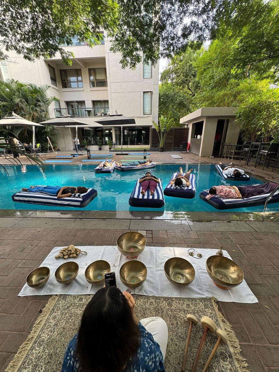 A practitioner seated before a row of brass singing bowls, guests floating in the pool beyond