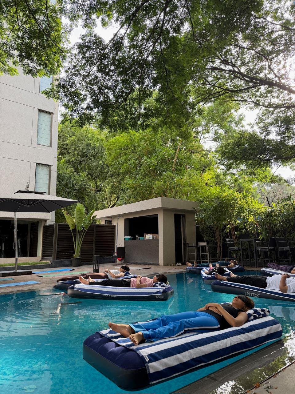 Guests lying on striped floating beds in a tree-shaded hotel pool, eyes closed