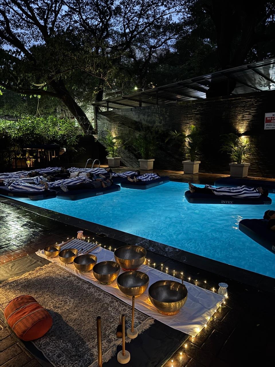 Guests resting on floating beds in a softly lit pool at dusk, brass singing bowls glowing by candlelight at the water's edge