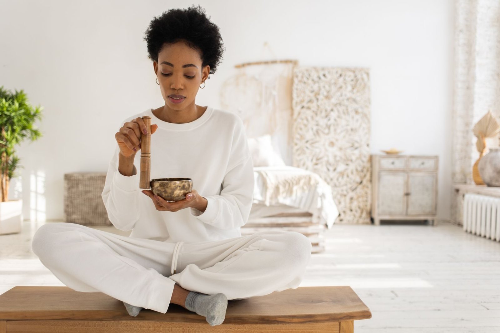 A woman seated cross-legged in a bright room, holding a singing bowl