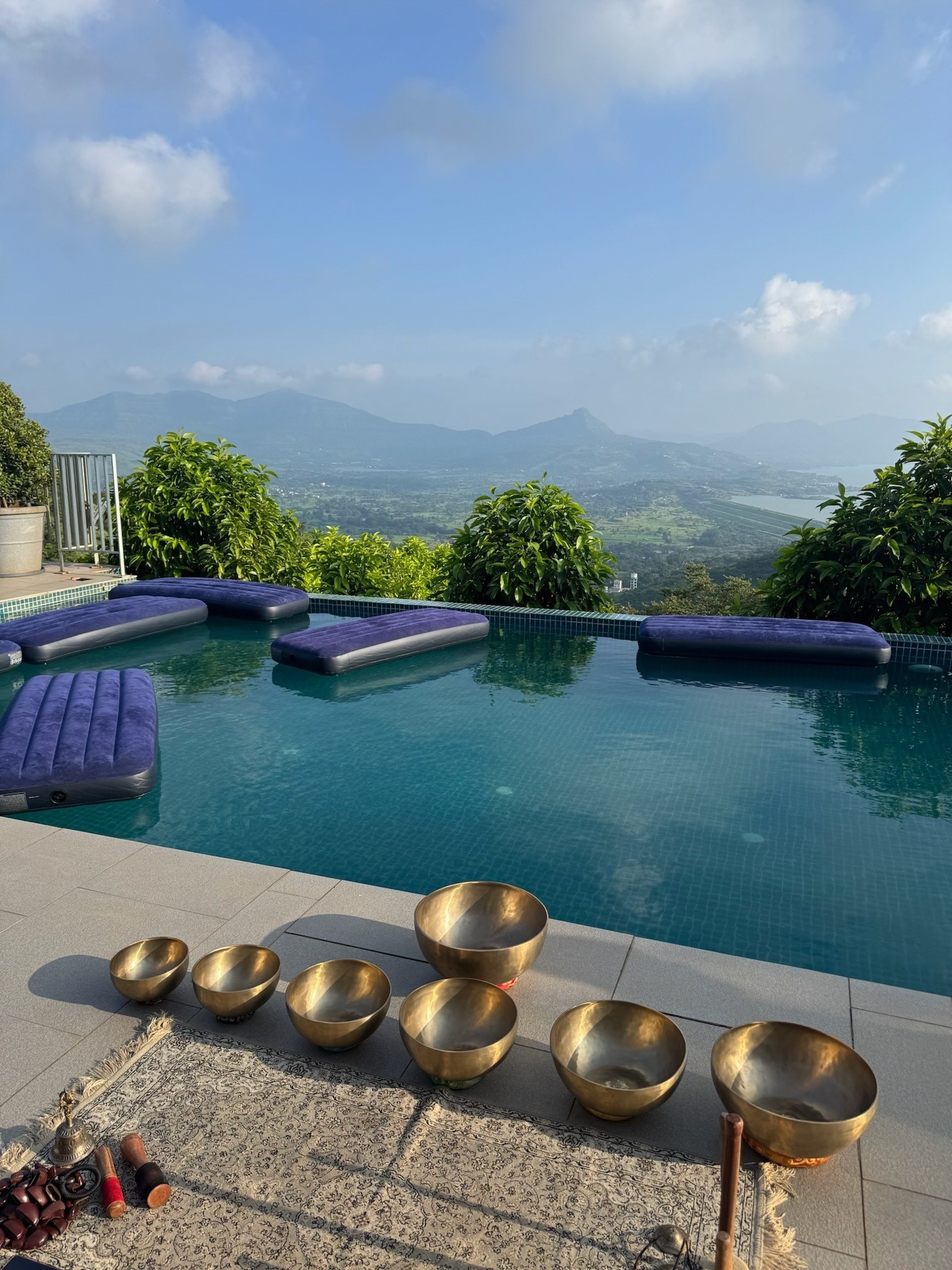 Brass singing bowls laid out on the deck of a hilltop infinity pool, floating beds on the water and green hills beyond